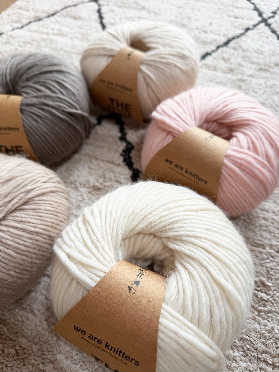 We Are Knitters Petite Wool yarn balls in cream, beige, gray and pink on a patterned rug.