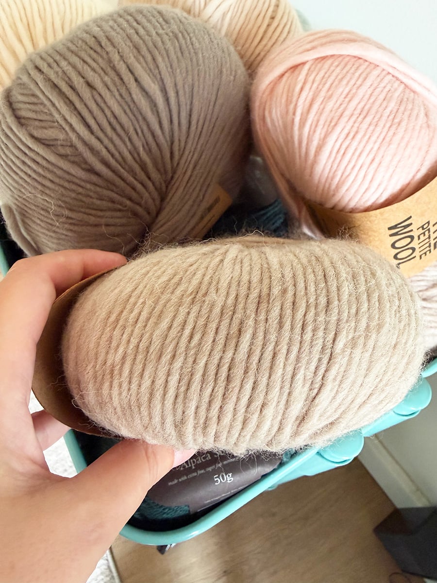 Hand holding a beige ball of bulky yarn beside gray, cream and pink yarn balls.